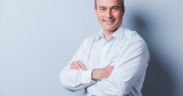 Confident mature man. Portrait of confident mature man in shirt looking at camera and smiling while keeping arms crossed and standing against grey background